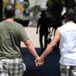 A picture of two men (Rudy Reyes, and Chris Hannifin) holding hands while running together. Behind waves an LGBTQ+ flag
